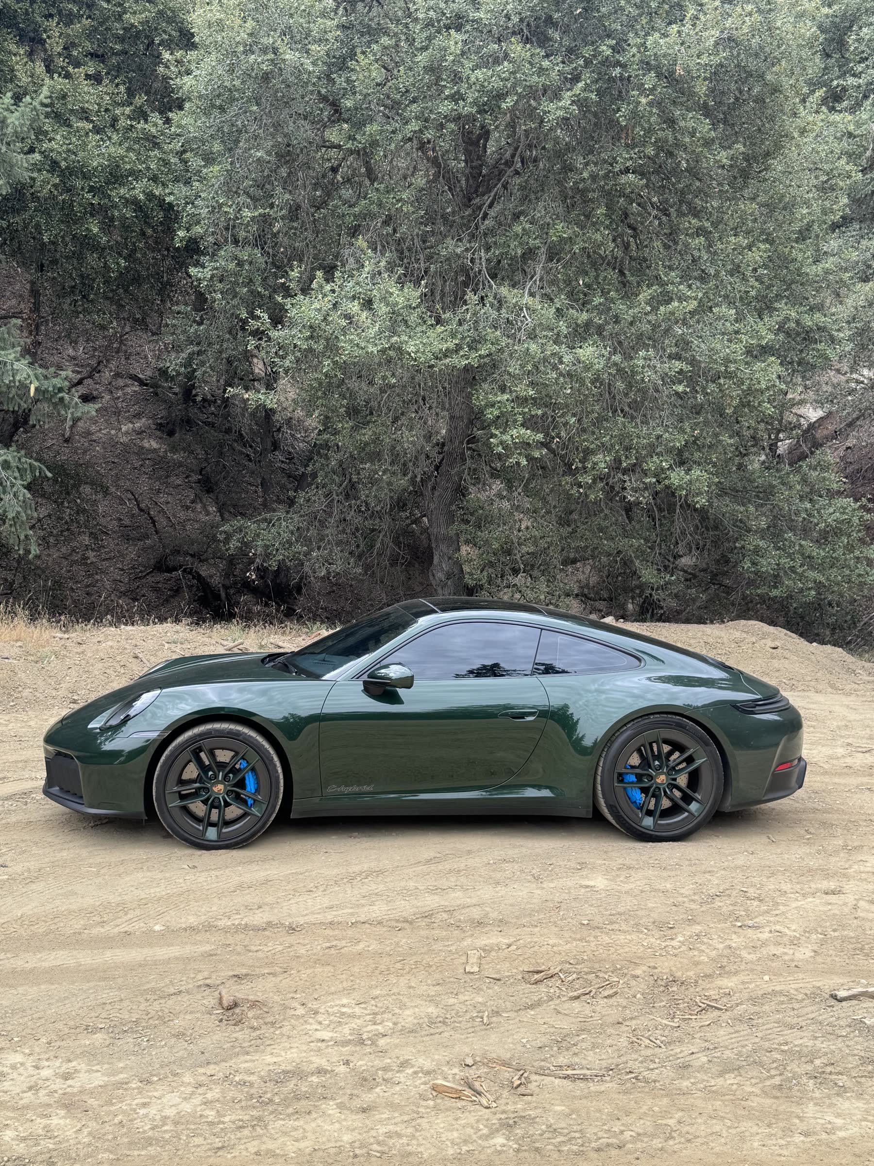 Side profile of an oak green Porsche 911 Carrera 4 GTS on a dirt road below a hillside of trees